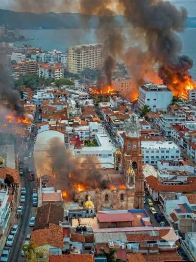 A street scene in Mexico showing the aftermath of cartel-related violence, with smoke rising from burning vehicles.